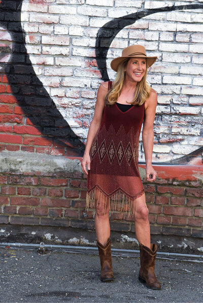 Kristy Clarke, owner of The WiLD Collective, stands in a dress and cowboy boots next to a textured wall.