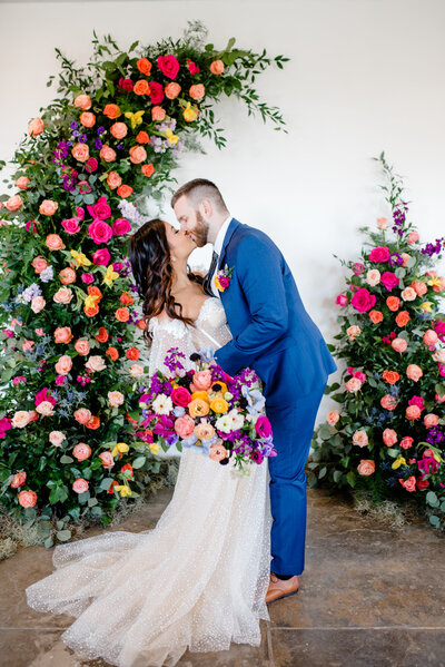 A bride and groom kissing with florals behind them.