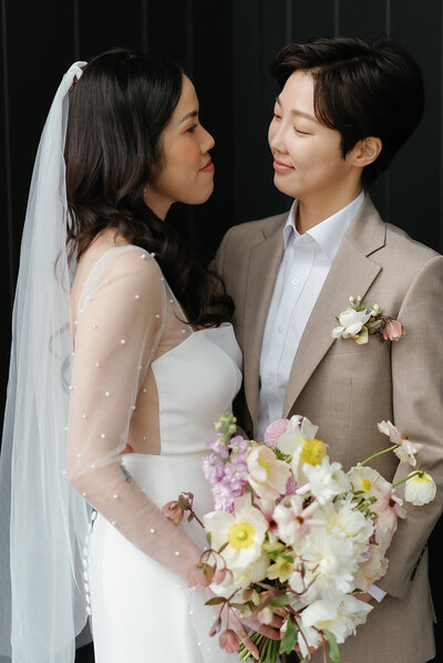 A bride in a veil holding a bouquet looks lovingly at her partner in a tan suit.