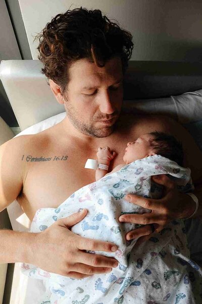 A dad gently holds his newborn baby in a hospital bed, with warm natural light streaming in from the window.