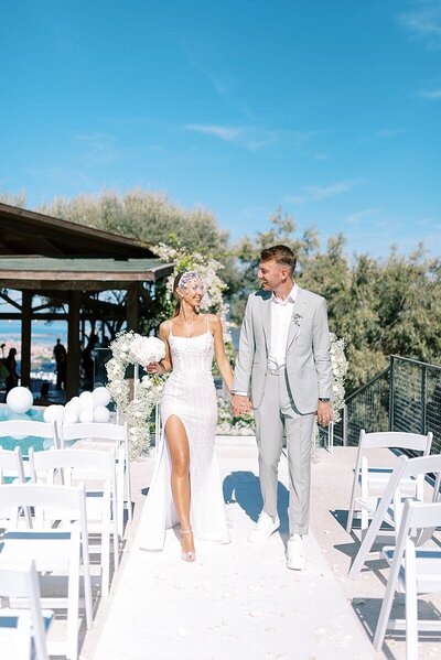 Bride and groom walk down the aisle in celebration at Villa Apanemia in Lefkada, with white floral decor and blue skies at their destination wedding
