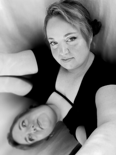 Black and while picture of a plus sized woman smiling into the camera with bangs framing her face and illuminated by the glow of sunshine reflected on her pale skin from a mirror she is holding