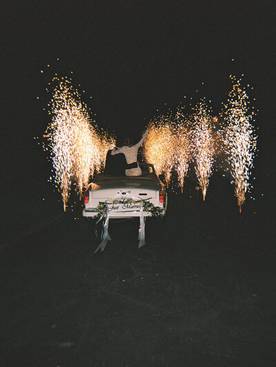 Photo from behind of bride and groom driving away from wedding in the dark with car surrounded by cold sparks