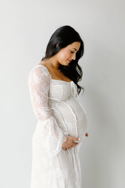 Expecting mother wearing white, lace dress and looking at baby bump during Indianapolis maternity photos