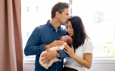 Loving parents hold and kiss their newborn in a bright, natural-light home setting, beautifully documented by Ellobelle Photography.