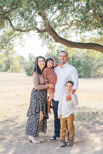 A family of four stand under a tree at Brushy Creek Lake Park. The father holds the daughter, the son stands to the side of his father, and the mother wraps her arms around her daughter. They all look at the camera smiling in this posed shot taken by an Austin Family Photographer