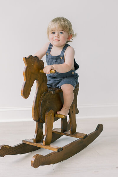 A toddler sitting proudly on a wooden rocking horse, looking toward the camera in soft studio light — Raleigh portrait photography.