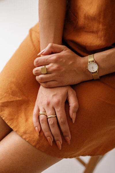 Woman's hand crossed in her lap wearing a gold watch and rings and an orange dress