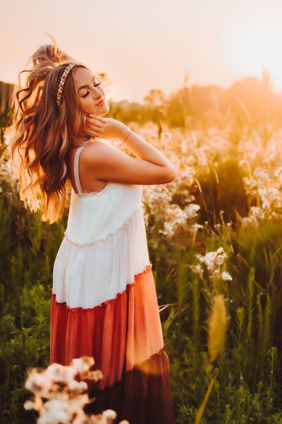 a young girl dressed in a white and red striped dress posing with her eyes closed in a field during her senior photoshoot