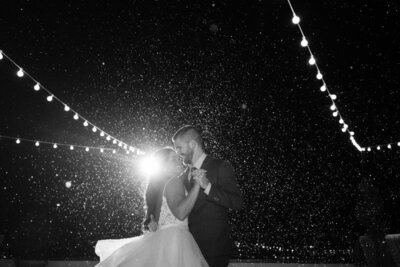 Black and white photograph of a bride and groom dancing intimately under twinkling string lights during a snowy evening at their wedding reception at the White Room in historic downtown St. Augustine. The couple is silhouetted against falling snow with dramatic backlighting creating a romantic, dreamy atmosphere.
