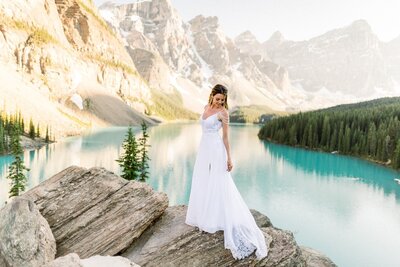 This stunning bridal portrait captures the bride gracefully seated on a rock at Moraine Lake in Banff National Park. The serene turquoise waters and towering mountain peaks create a breathtaking backdrop, perfectly complementing the bride's elegance and the natural beauty of the moment.