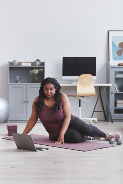 A curvy Black woman sitting on a yoga mat next to a laptop
