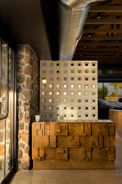Modern restaurant entrance with stone wall, textured wooden reception desk, and decorative perforated divider panel. 