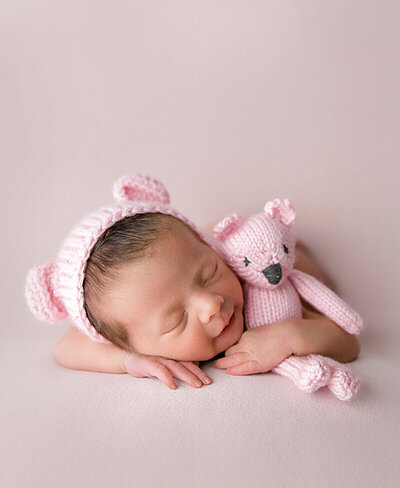 A newborn baby smiles and holds a little bear during her posed newborn session in the studio.