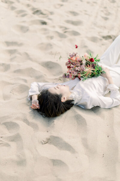 Kohnur minimalist bride with natural makeup and short textured hair, wearing a white long-sleeved top and lying on the sandy beach with a bouquet.