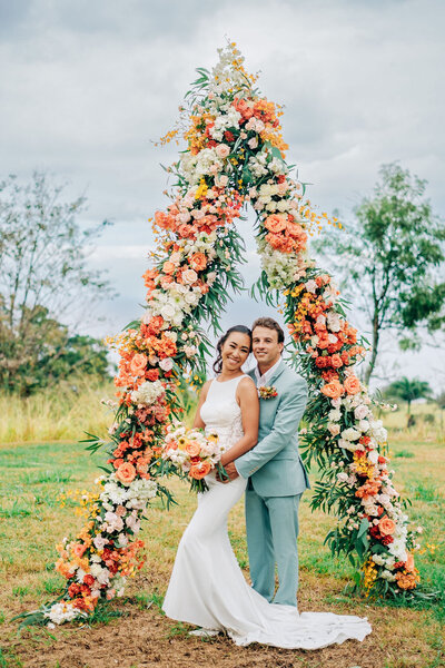 Stunning triangle wedding arch overlooking the Pacific Ocean at Puuwai Gardens on Hawaii Island. Adorned with white, yellow, persimmon, and orange flowers, this breathtaking ceremony setup embodies tropical elegance for a luxury destination wedding.
