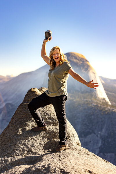 Female photographer standing on cliff in Yosemite Park holding camera with hands out.