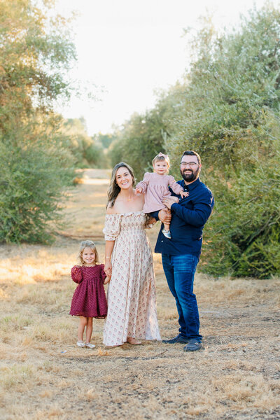 Happy family of four standing in an olive orchard with golden light during a photo session with One Good Shot Photography family photographer