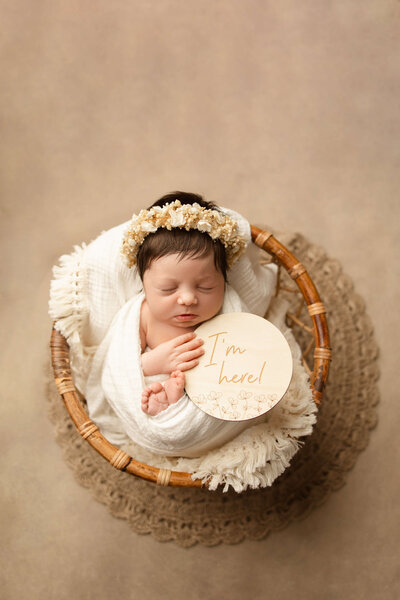 Newborn baby boy in a blue swaddle in a tan basket on a tan background