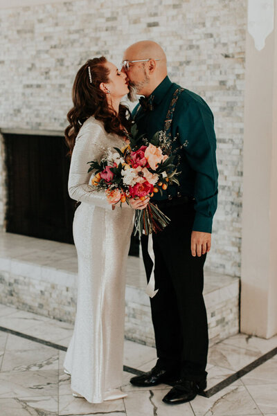 A newlywed couple shares an intimate kiss, with the bride in a white gown holding a vibrant bouquet. Wedding coordinated by WINK! Weddings in California.