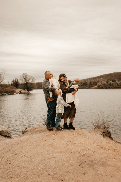 Family of five standing together on a lakeside rock during an outdoor photo session in Stanhope, NJ, with autumn trees and hills in the background, captured by Burkat Photography