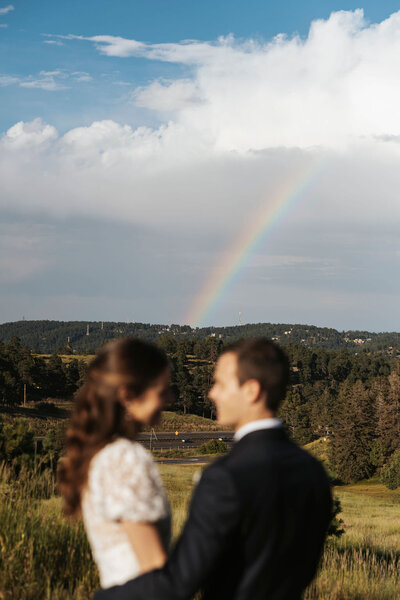 A newlywed couple standing close looking at each other with a rainbow in the distance.