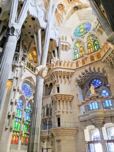 Interior of the Sagrada Familia in Barcelona, featuring tall columns and vibrant stained glass windows with intricate designs, casting colorful light onto the stone walls and architectural details.