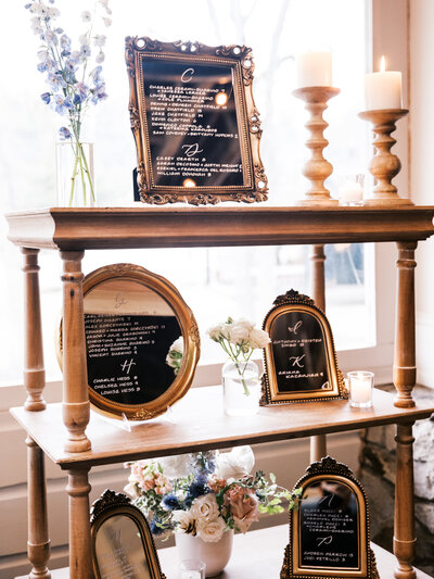 Vintage gold mirror seating chart display with calligraphy on wooden shelf with florals for wedding at Cedar Lakes Estate in Hudson Valley, New York.
