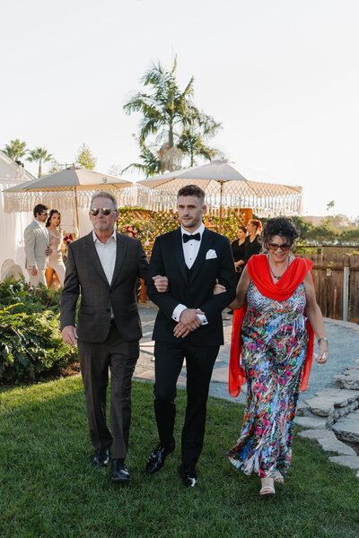 A groom in a black tuxedo walks arm-in-arm with his parents during a sunny outdoor wedding ceremony.