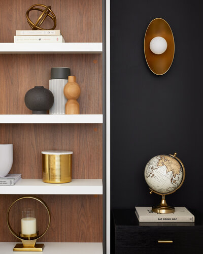 White shelving with wood backing next to a black wall containing various art pieces with bronze accents