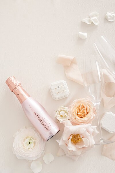 Champagne bottle & glasses styled with blooms, petals, ribbon, and wedding rings in a ring box.