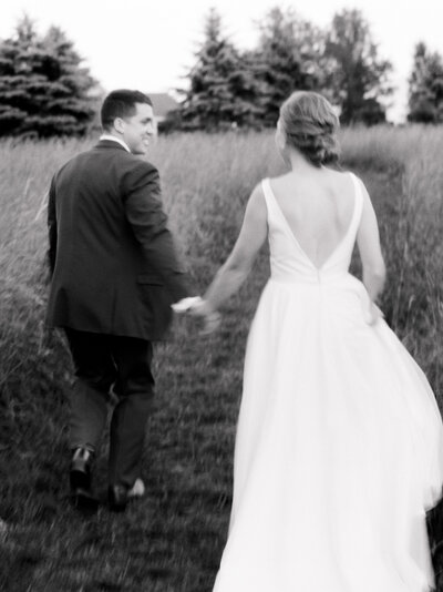Black and white photo of a bride and groom holding hands and walking away from the camera in a grassy field