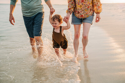 mom and dad holding hands with child running down beach