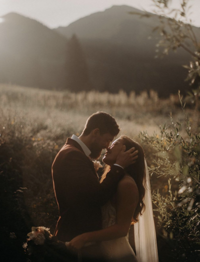 A bride and groom kissing in a field at the foot of a mountain. The groom has his hand place on the side of the brides face