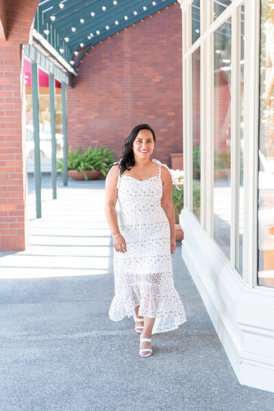 Woman in white dress walking toward camera next to a store front window during her brand photography session.