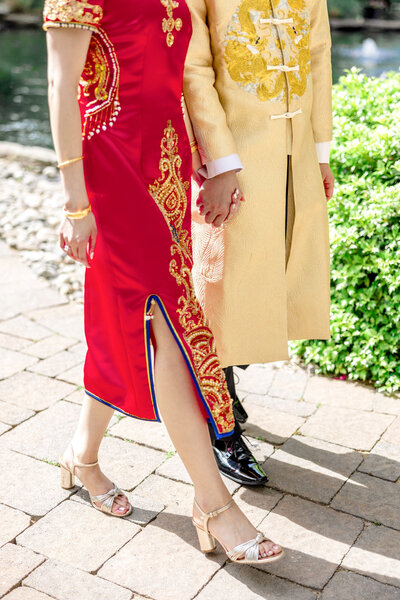 A couple in traditional attire holds hands while walking. The woman wears a red dress with gold embroidery, and the man wears a gold embroidered outfit. They are on a stone pathway with greenery and water in the background.