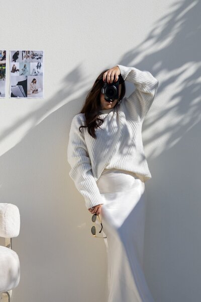 A woman stands against a white wall bathed in sunlight, dressed in a flowing white satin skirt and a cozy white knitted sweater. She holds a camera to her eye with one hand, while sunglasses rest in the other, embodying a serene yet adventurous vibe.