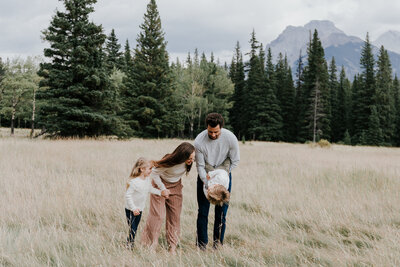 A family enjoys time together in a field with pine trees and  mountains in the background. The father is tickling his son while the mother leans over and smiles at her son and holds her daughters hand.