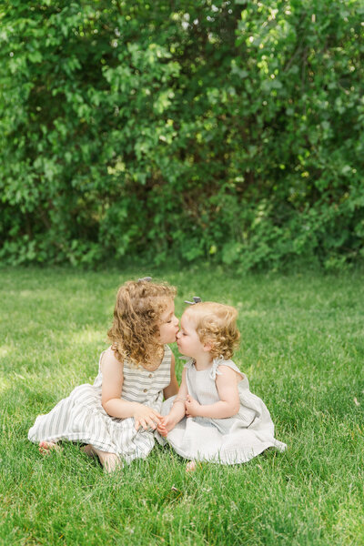 siblings sitting in an outdoor setting, the older sister kisses the younger sister on the forehead