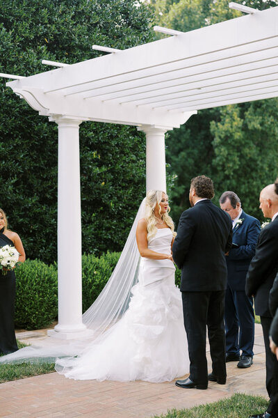 Kailea and Zach holding hands under an archway on the lawn of Boxwood Estate. During their ceremony, Kailea is wearing a white gown with white veil while Zach is wearing a classic suit. 