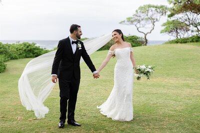 A newlywed couple holding hands and walking together as one of their veils flows behind them 