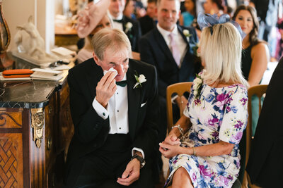 During an indoor ceremony at Pylewell Park, an older man in a tuxedo wipes a tear. A woman in a floral dress sits beside him, smiling warmly. In the background, other guests are seated, dressed in formal attire.
