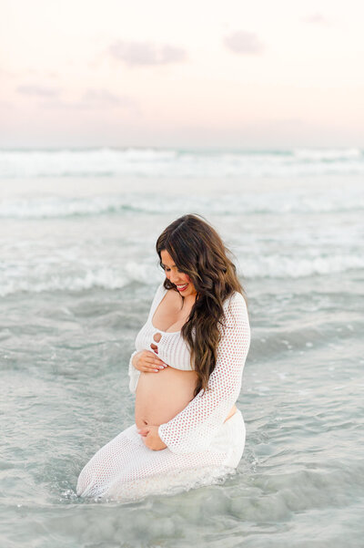 Orlando maternity photographer captures pregnant mom sitting in ocean holding her belly at sunset