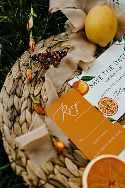 Orange, lemons, orange flowers, monarch butterflies in a woven basket with a Save the Date card and RSVP card present.