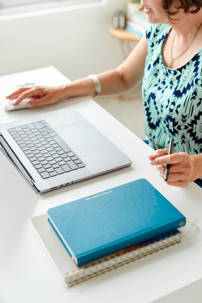Kate Daniel, a Calgary brand photographer, works at her desk with a laptop, using a mouse while holding a pen in her other hand. A blue ‘Full Focus Planner’ sits on top of a spiral notebook, adding a sense of organization and productivity. The bright, modern workspace is complemented by a small side table with stacked books and a decorative element in the background.