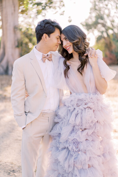 Portrait of a bride and groom wearing a khaki suit, bow tie and a lavender gown leaning into each other smiling outdoors