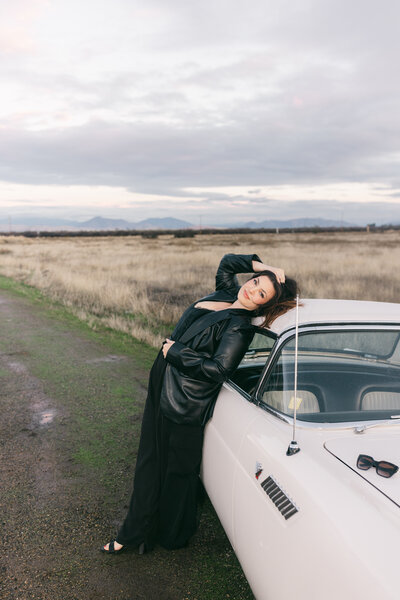 Woman leaning against a vintage white car on a rural road at sunset in Greenville, South Carolina, wearing a black leather jacket and trousers. Editorial portrait photography by Dani Lacey Photographs.