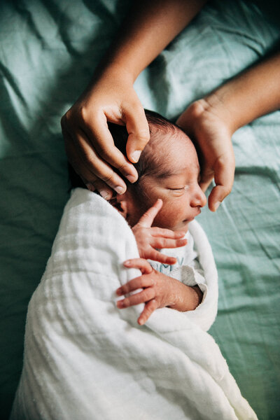 newborn baby boy in white swaddle blanket with his head cradled by his older brother's hands