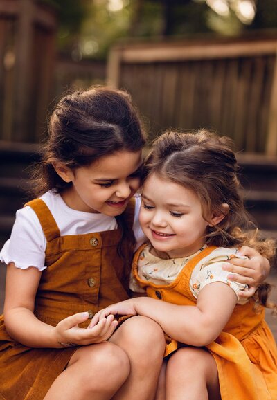Two young girls sitting closely on a bench, sharing a moment of friendship and joy in a sunny outdoor setting.