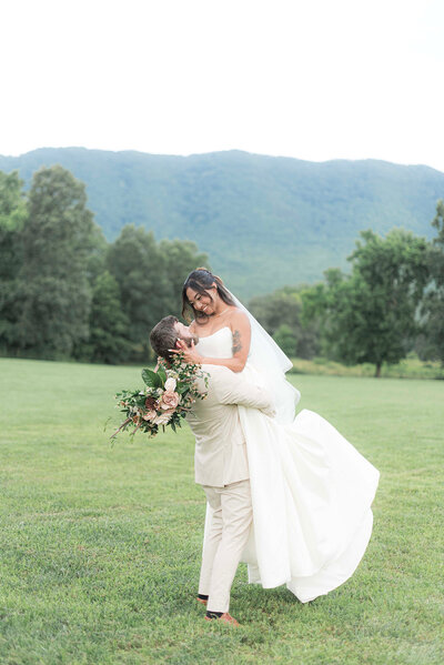 Groom in a cream-colored tuxedo lifts his bride in the air in front of a beautiful mountain scenic background